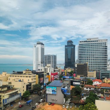 View over Colombo, Sri Lanka: high-rise office and hotel towers beside the sea, with low-rise tiled-roof neighbourhoods spreading inland.