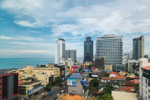 View over Colombo, Sri Lanka: high-rise office and hotel towers beside the sea, with low-rise tiled-roof neighbourhoods spreading inland.